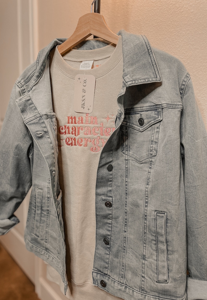 May include: A light gray denim jacket with a light gray sweatshirt underneath. The sweatshirt has the text "main character energy" in pink lettering. A white tag with the text "JKSNY & CO!" is attached to the sweatshirt.