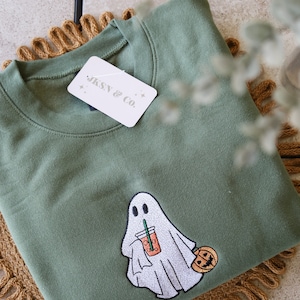 May include: A green sweatshirt with an embroidered ghost holding a pumpkin spice latte and a small pumpkin. The sweatshirt has a white tag with the text "JKSN & CO."