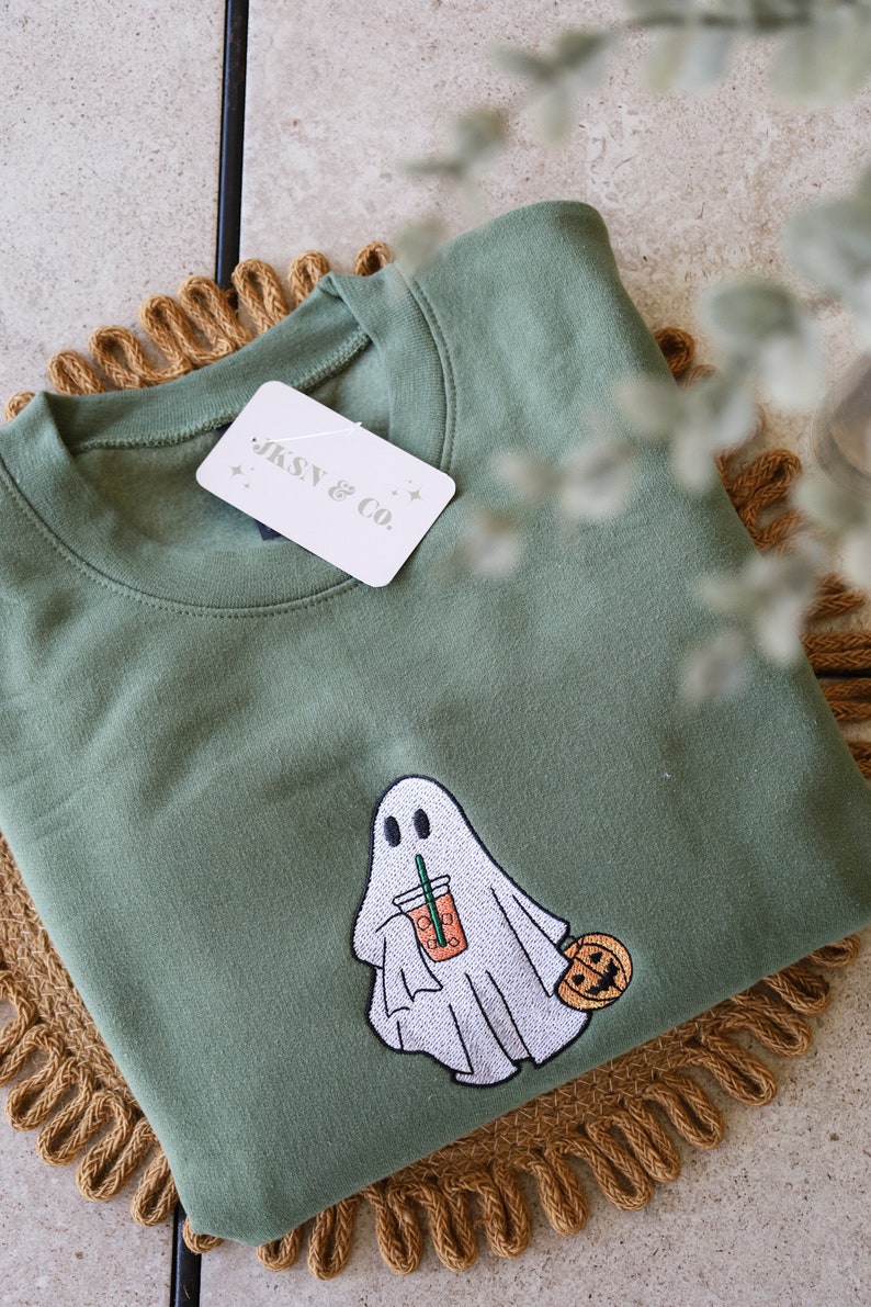 May include: A green sweatshirt with an embroidered ghost holding a pumpkin spice latte and a small pumpkin. The sweatshirt has a white tag with the text "JKSN & CO."