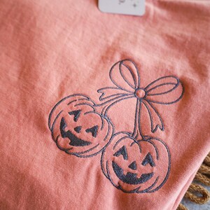 Op de afbeelding: Koraalrood T-shirt met een geborduurd ontwerp van twee lachende pompoenen, verbonden door een strik. Een label met de tekst "JKXY & CO." is aan het shirt bevestigd.