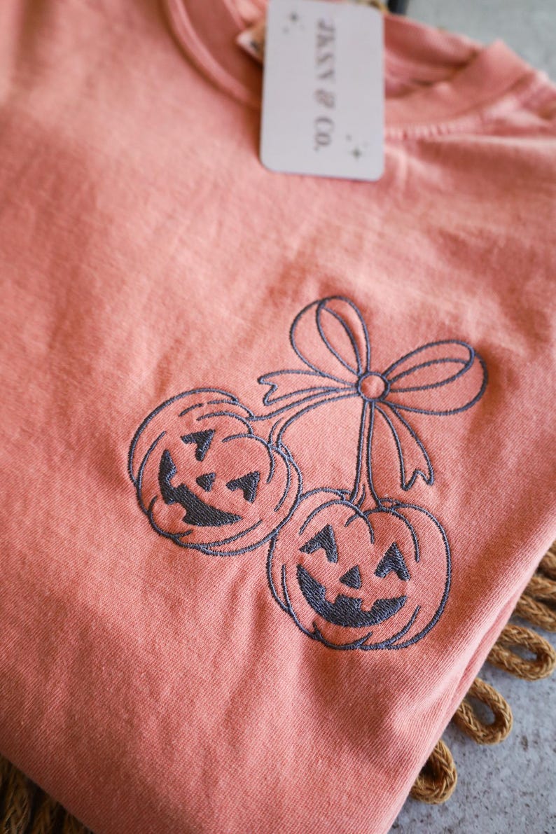 Op de afbeelding: Koraalrood T-shirt met een geborduurd ontwerp van twee lachende pompoenen, verbonden door een strik. Een label met de tekst "JKXY & CO." is aan het shirt bevestigd.