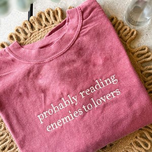 May include: A dusty rose colored t-shirt with the text "probably reading enemies to lovers" embroidered in white.