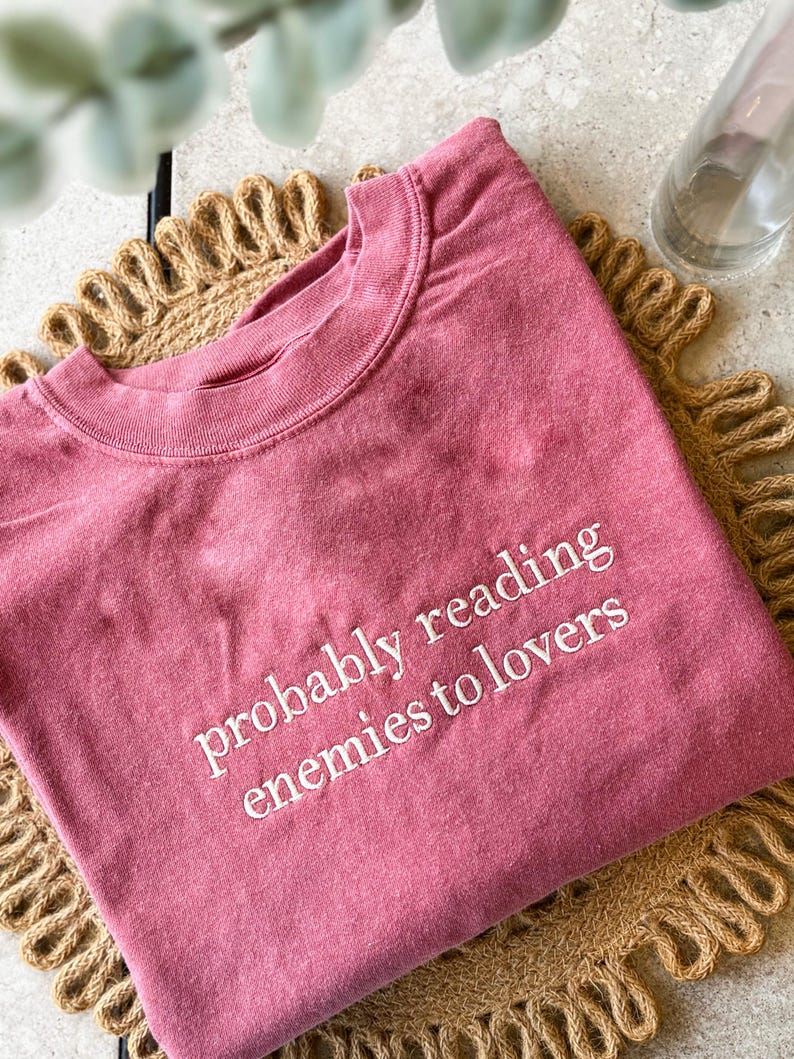 May include: A dusty rose colored t-shirt with the text "probably reading enemies to lovers" embroidered in white.
