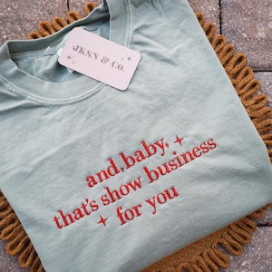 Puede incluir: Una camiseta verde claro con texto bordado en rojo que dice "and, baby, that's show business + for you". La camiseta tiene una etiqueta que dice "JKSN & Co." y est&aacute; sobre una superficie marr&oacute;n texturizada.