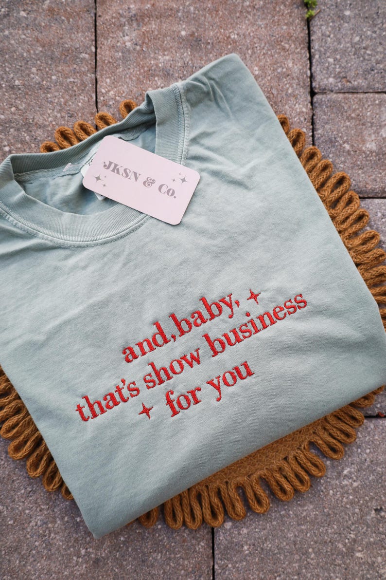 Puede incluir: Una camiseta verde claro con texto bordado en rojo que dice "and, baby, that's show business + for you". La camiseta tiene una etiqueta que dice "JKSN & Co." y est&aacute; sobre una superficie marr&oacute;n texturizada.