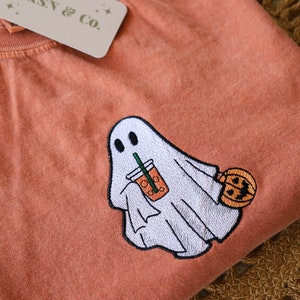 Może przedstawiać: Pomarańczowy t-shirt z haftowanym duchem trzymającym kubek z lodowatą kawą i dynią. Na koszulce znajduje się mała metka z napisem "JKSN & Co."