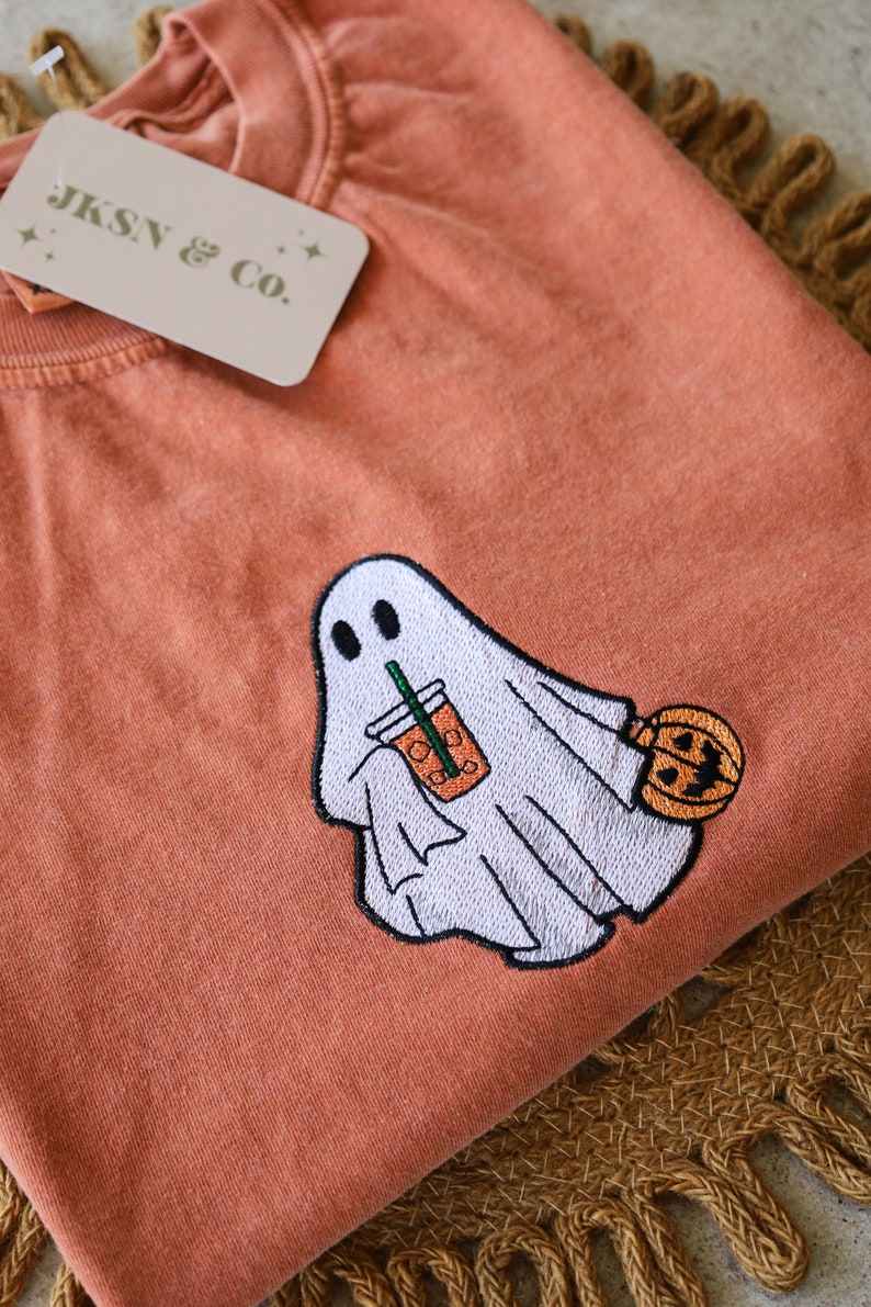 Może przedstawiać: Pomarańczowy t-shirt z haftowanym duchem trzymającym kubek z lodowatą kawą i dynią. Na koszulce znajduje się mała metka z napisem "JKSN & Co."