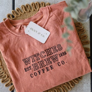 Puede incluir: Una camiseta de color naranja quemado con el texto "WITCHES BREW EST. 1626 COFFEE CO." en bordado negro. La camiseta est&aacute; doblada y sobre una estera tejida de color marr&oacute;n.