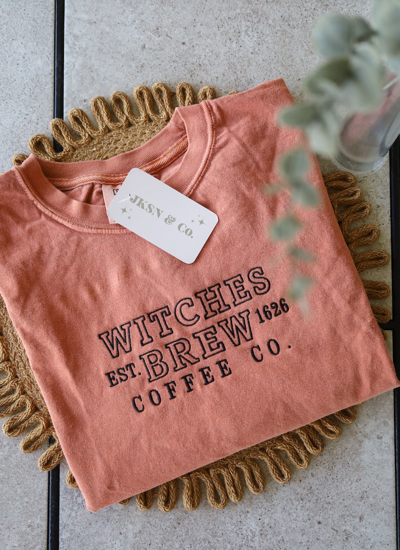 Puede incluir: Una camiseta de color naranja quemado con el texto "WITCHES BREW EST. 1626 COFFEE CO." en bordado negro. La camiseta est&aacute; doblada y sobre una estera tejida de color marr&oacute;n.