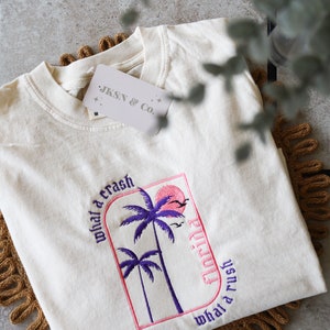 May include: A white t-shirt with a pink and purple embroidered design featuring palm trees and the text "what a crash Florida what a rush". The shirt also has a small white tag with the text "JKSN & CO."