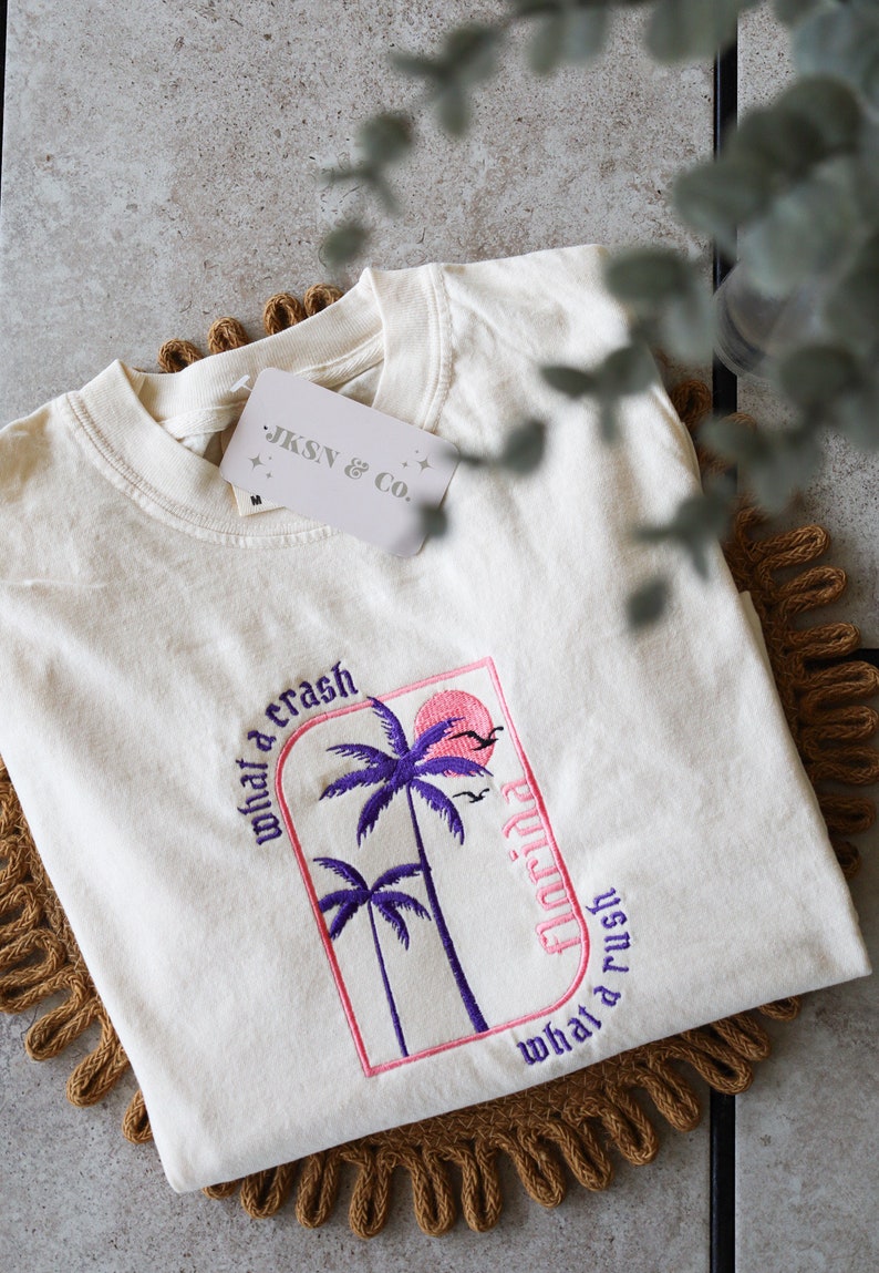 May include: A white t-shirt with a pink and purple embroidered design featuring palm trees and the text "what a crash Florida what a rush". The shirt also has a small white tag with the text "JKSN & CO."