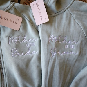 May include: Two sage green zip-up hoodies featuring white embroidered text. One hoodie reads Mother of the Bride and the other reads Mother of the Groom. Each garment includes a light pink tag with the text JKSN & Co. and star icons.