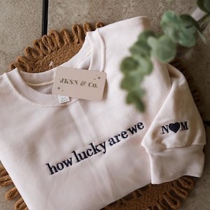 May include: A cream colored sweatshirt with the text "how lucky are we" embroidered in navy blue on the front. The left sleeve has the text "N♥M" embroidered in navy blue.