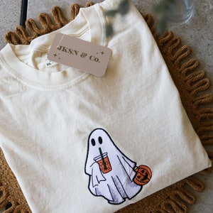 Może przedstawiać: Biały T-shirt z haftowanym duchem trzymającym dynię i nap&oacute;j. Koszulka ma metkę z napisem "JKSN & Co."