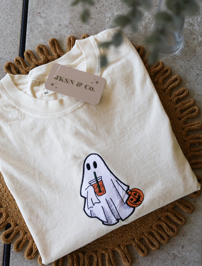 Może przedstawiać: Biały T-shirt z haftowanym duchem trzymającym dynię i nap&oacute;j. Koszulka ma metkę z napisem "JKSN & Co."
