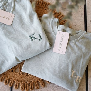 May include: Two light blue t-shirts with embroidered initials. The t-shirts are folded and placed on a brown woven mat. A white tag with the text "JKSN & CO." is attached to one of the t-shirts.