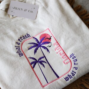 May include: A white t-shirt with an embroidered design featuring palm trees, a pink sun, and the text "what a crash" and "what a rush" in a gothic font. The text "Playa" is also embroidered in a gothic font.