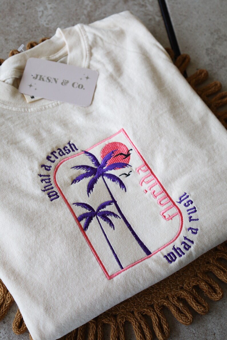 May include: A white t-shirt with an embroidered design featuring palm trees, a pink sun, and the text "what a crash" and "what a rush" in a gothic font. The text "Playa" is also embroidered in a gothic font.
