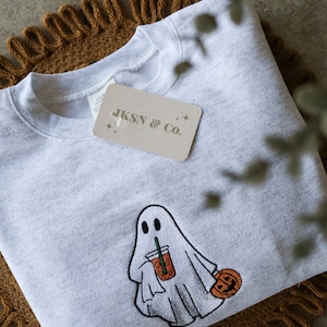 May include: A gray sweatshirt with an embroidered design of a ghost holding a pumpkin spice latte and a small pumpkin. The sweatshirt has a tag that says "JKSN & Co."