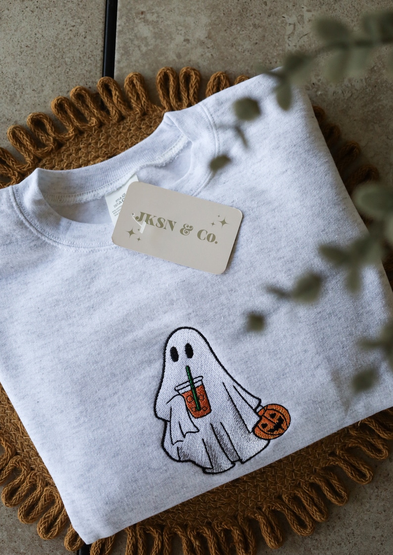 May include: A gray sweatshirt with an embroidered design of a ghost holding a pumpkin spice latte and a small pumpkin. The sweatshirt has a tag that says "JKSN & Co."