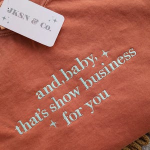 Puede incluir: Camiseta de color &oacute;xido con la frase bordada en azul claro "and, baby, that's show business for you". Una etiqueta blanca con "JKSN & Co." est&aacute; adjunta. La camiseta est&aacute; doblada sobre una superficie texturizada.