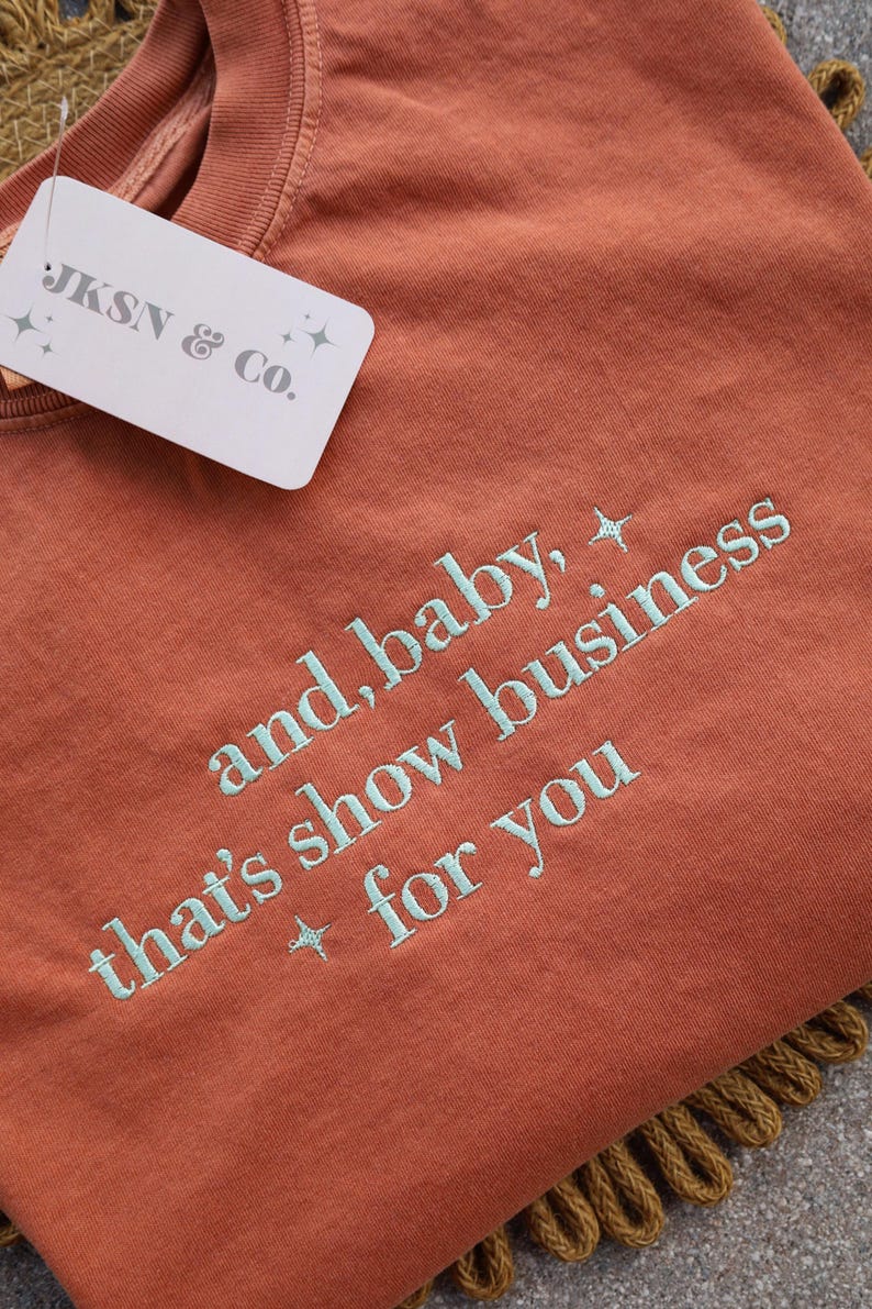 Puede incluir: Camiseta de color &oacute;xido con la frase bordada en azul claro "and, baby, that's show business for you". Una etiqueta blanca con "JKSN & Co." est&aacute; adjunta. La camiseta est&aacute; doblada sobre una superficie texturizada.