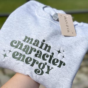 May include: Gray sweatshirt with the text "main character energy" embroidered in green. The text is in a retro font style.