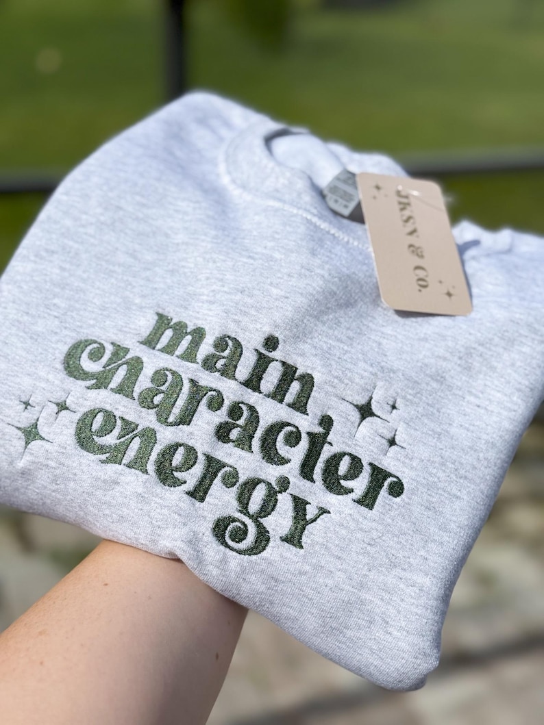 May include: Gray sweatshirt with the text "main character energy" embroidered in green. The text is in a retro font style.