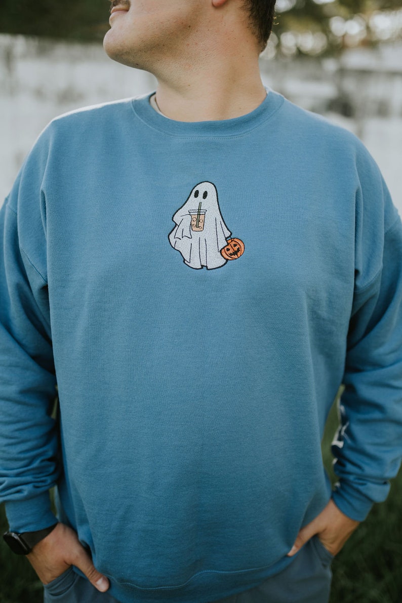 May include: A blue sweatshirt with a white ghost holding a pumpkin and a cup of coffee. The ghost has a friendly face and is wearing a white sheet.