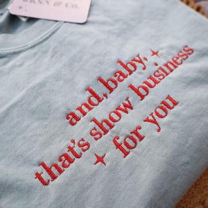 Puede incluir: Camiseta azul claro con texto bordado en rojo: "and, baby, that's show business for you." Una etiqueta "JKNY & CO." est&aacute; adjunta. La camiseta est&aacute; doblada sobre una superficie marr&oacute;n.