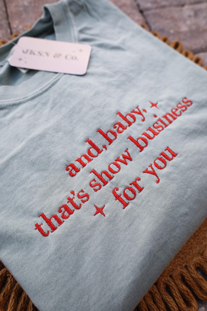 Puede incluir: Camiseta azul claro con texto bordado en rojo: "and, baby, that's show business for you." Una etiqueta "JKNY & CO." est&aacute; adjunta. La camiseta est&aacute; doblada sobre una superficie marr&oacute;n.