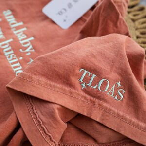 Puede incluir: Una camiseta de color terracota con el texto "and baby, it's my business" impreso en azul claro. El pu&ntilde;o de la manga presenta las letras bordadas "TLOA'S" en azul claro. Una etiqueta blanca est&aacute; adjunta.