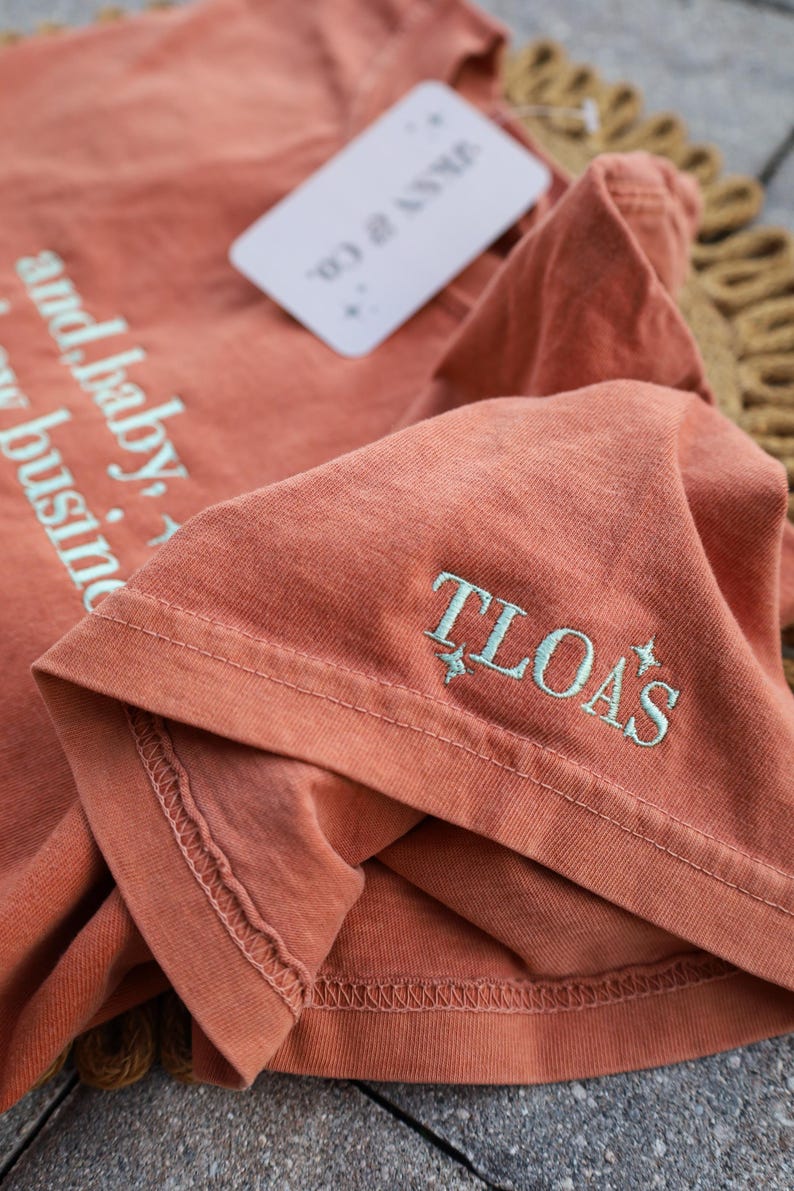 Puede incluir: Una camiseta de color terracota con el texto "and baby, it's my business" impreso en azul claro. El pu&ntilde;o de la manga presenta las letras bordadas "TLOA'S" en azul claro. Una etiqueta blanca est&aacute; adjunta.