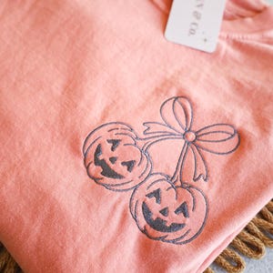 Op de afbeelding: Koraalrood T-shirt met een geborduurd ontwerp van twee lachende pompoenen en een strik. Het shirt heeft een label met de tekst "JENNY & CO.". Een feestelijke top voor Halloween.
