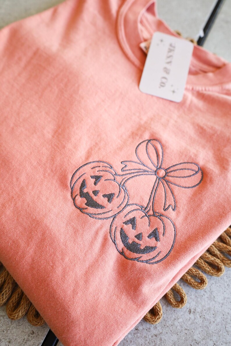 Op de afbeelding: Koraalrood T-shirt met een geborduurd ontwerp van twee lachende pompoenen en een strik. Het shirt heeft een label met de tekst "JENNY & CO.". Een feestelijke top voor Halloween.