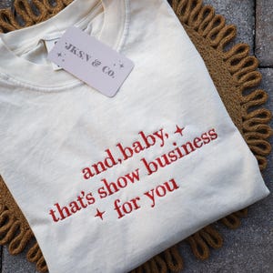 Puede incluir: Camiseta color crema con texto bordado en rojo: "and, baby, that's show business + for you". La camiseta tiene cuello redondo y una etiqueta que dice "JKSN & Co.". Se muestra sobre una estera tejida marr&oacute;n y circular.