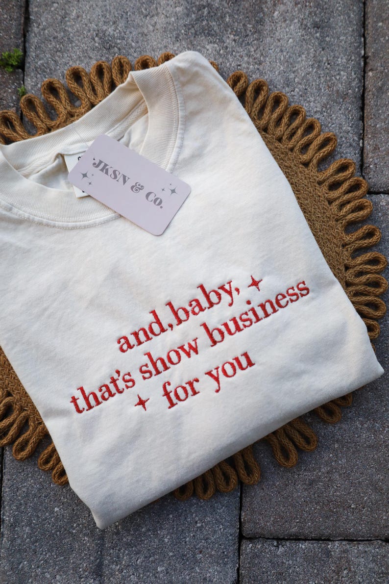 Puede incluir: Camiseta color crema con texto bordado en rojo: "and, baby, that's show business + for you". La camiseta tiene cuello redondo y una etiqueta que dice "JKSN & Co.". Se muestra sobre una estera tejida marr&oacute;n y circular.