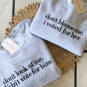 May include: Two gray sweatshirts with black embroidered text. The top sweatshirt says "don't blame me, i voted for her." The bottom sweatshirt says "don't look at me, i didn't vote for him."