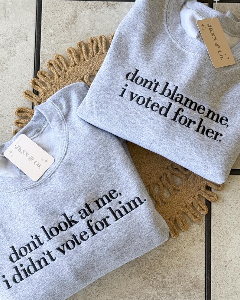 May include: Two gray sweatshirts with black embroidered text. The top sweatshirt says "don't blame me, i voted for her." The bottom sweatshirt says "don't look at me, i didn't vote for him."