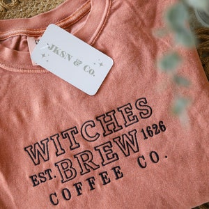 Puede incluir: Una camiseta de color melocot&oacute;n con el texto "WITCHES BREW COFFEE CO. EST. 1626" bordado en negro. Una etiqueta blanca con el texto "JKSN & CO." est&aacute; unida a la camiseta.