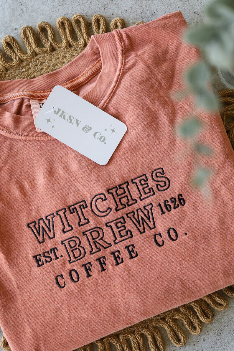 Puede incluir: Una camiseta de color melocot&oacute;n con el texto "WITCHES BREW COFFEE CO. EST. 1626" bordado en negro. Una etiqueta blanca con el texto "JKSN & CO." est&aacute; unida a la camiseta.
