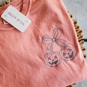 Op de afbeelding: Een koraalkleurig T-shirt met een ronde hals en een label met de tekst "JKSN & CO." heeft een geborduurd ontwerp van twee lachende pompoenen met een strik. Het shirt is op een bruin, getextureerd oppervlak gevouwen.