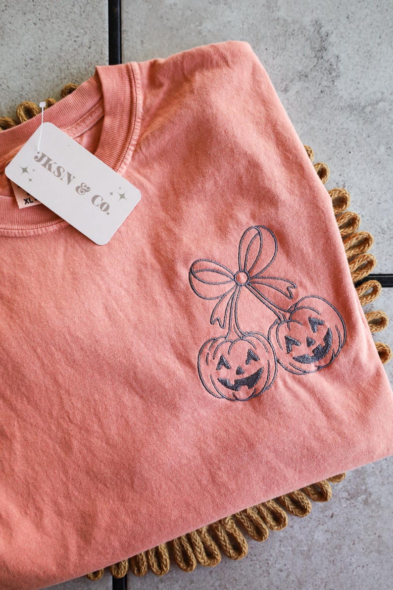 Op de afbeelding: Een koraalkleurig T-shirt met een ronde hals en een label met de tekst "JKSN & CO." heeft een geborduurd ontwerp van twee lachende pompoenen met een strik. Het shirt is op een bruin, getextureerd oppervlak gevouwen.