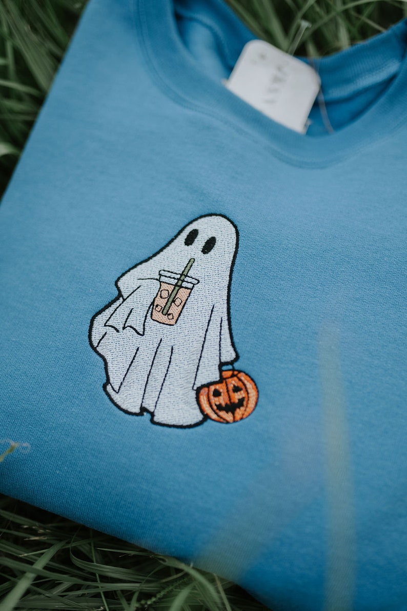 May include: A light blue sweatshirt with an embroidered ghost holding a pumpkin and a cup with a straw. The ghost is white and the pumpkin is orange.