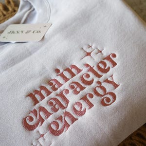 May include: A light gray sweatshirt with the embroidered phrase "main character energy" in a reddish-pink color. The sweatshirt has a tag that reads "JKSN & CO."