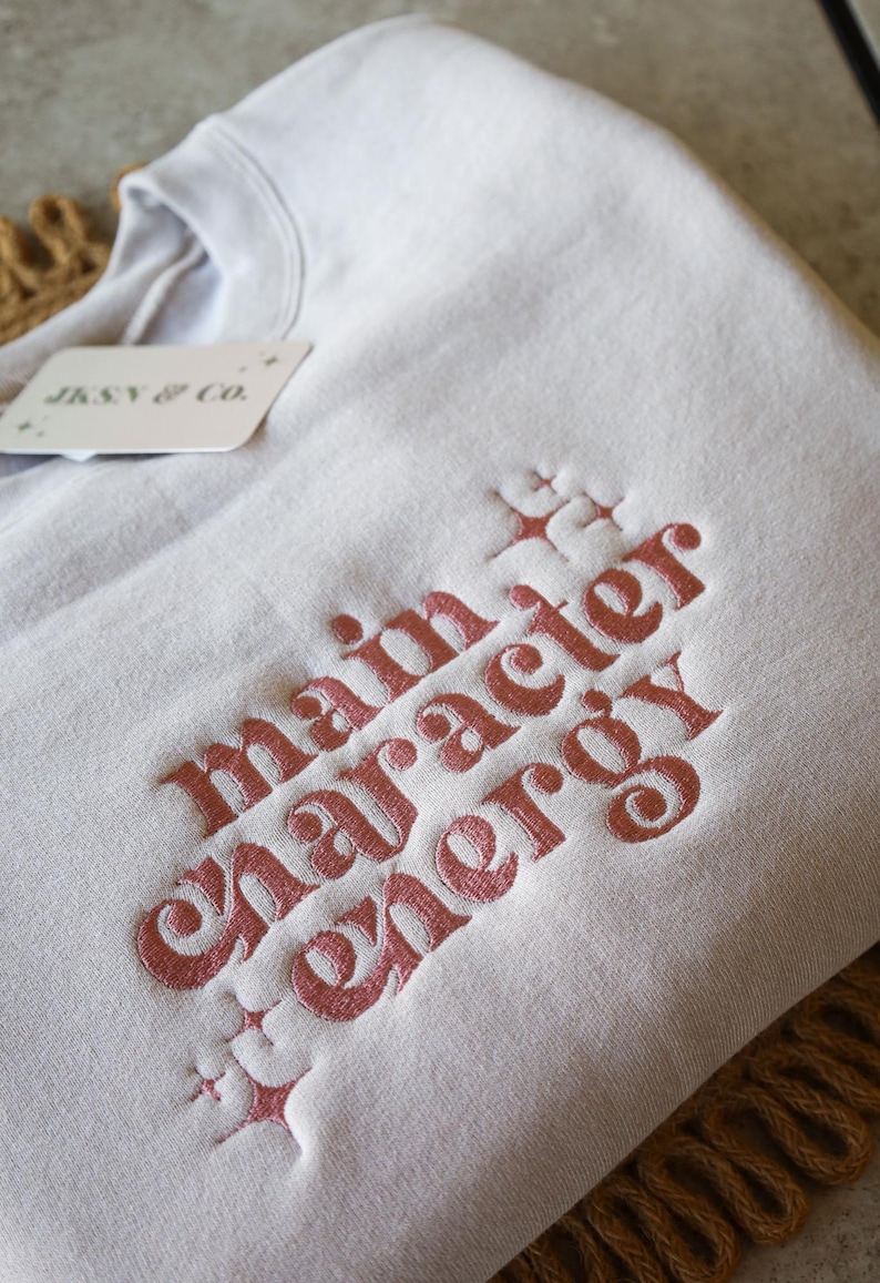 May include: A light gray sweatshirt with the embroidered phrase "main character energy" in a reddish-pink color. The sweatshirt has a tag that reads "JKSN & CO."