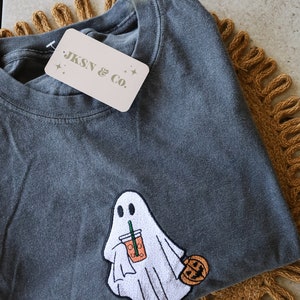 Może przedstawiać: Szary t-shirt z haftowanym duchem trzymającym latte z przyprawą dyniową i małą dynią. Na koszulce znajduje się metka z napisem "JKSN & Co."