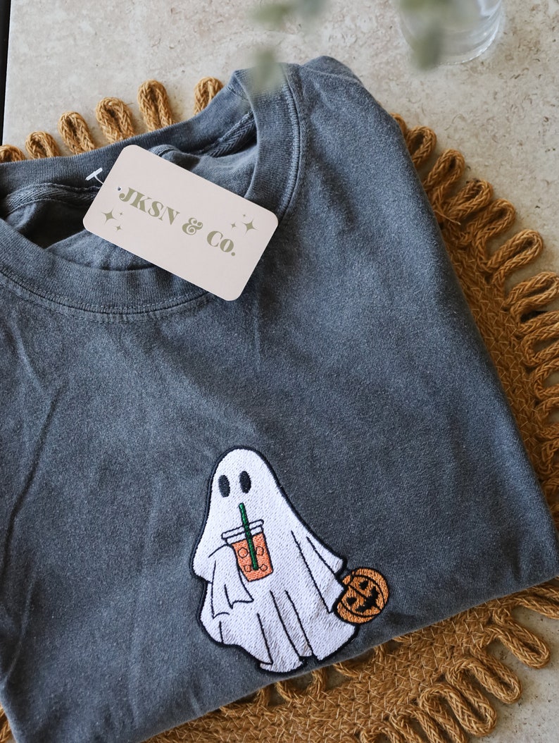 Może przedstawiać: Szary t-shirt z haftowanym duchem trzymającym latte z przyprawą dyniową i małą dynią. Na koszulce znajduje się metka z napisem "JKSN & Co."