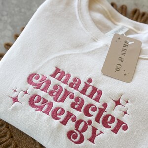 May include: A white sweatshirt with the phrase "main character energy" embroidered in pink. The sweatshirt has a tag that says "JAX & CO."