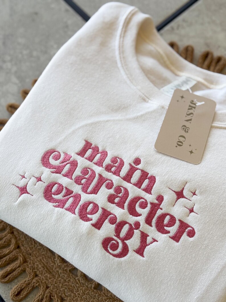 May include: A white sweatshirt with the phrase "main character energy" embroidered in pink. The sweatshirt has a tag that says "JAX & CO."